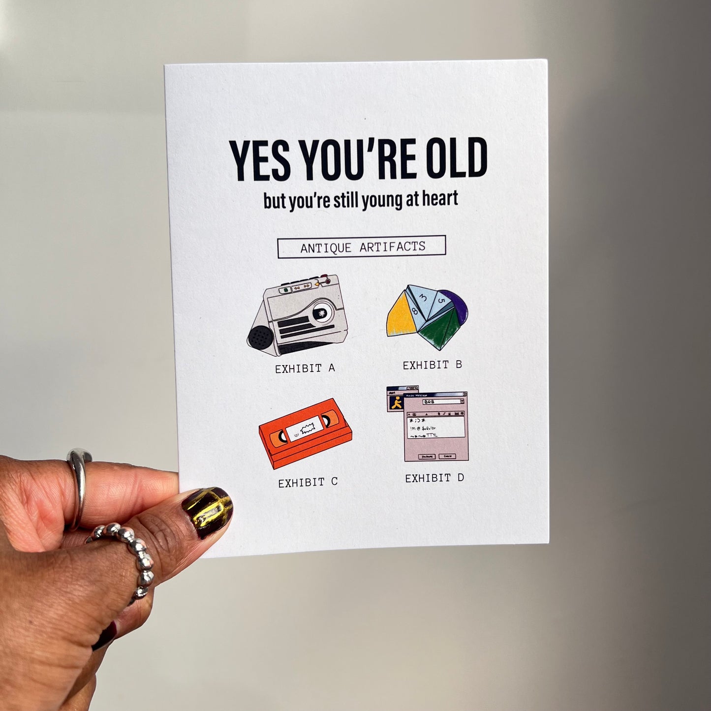 4.25 x 5.5” A2 size funny millennial birthday card with illustrations of a rugrats vhs, AIM screen, audio recorder, and paper fortune teller. The text reads, "Yes you’re old but you’re still young at heart". Message inside included. Black Woman Owned.