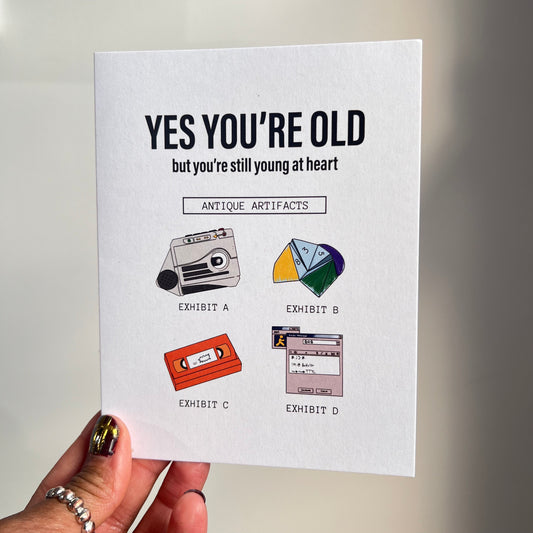 4.25 x 5.5” A2 size funny millennial birthday card with illustrations of a rugrats vhs, AIM screen, audio recorder, and paper fortune teller. The text reads, "Yes you’re old but you’re still young at heart". Message inside included. Black Woman Owned.