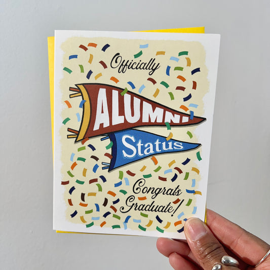 A2 size vibrant graduation card featuring colorful confetti and collegiate pennant banners. “Officially Alumni Status, Congrats Graduate!” text on the front with "Alumni” on the top burgundy banner and “Status!" below on a blue banner. Colorful confetti and a message inside the card. Envelope included.