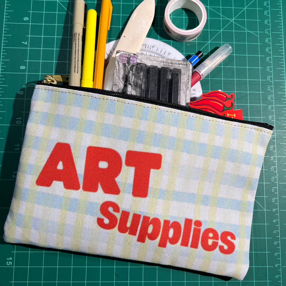 Two-sided polyester zip pouch with “Art Supplies” in a persimmon color with a white base and light blue and green gingham pattern. Includes a black zipper and gold zipper pull.