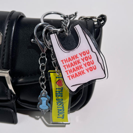 2.5” double-sided clear acrylic keychain featuring an illustrated white takeout bag with vibrant orange-red “thank you” text repeated on the bag. Comes with a lobster clasp, split ring, and chain. Bag charm for totes, crossbody bags, and purses.