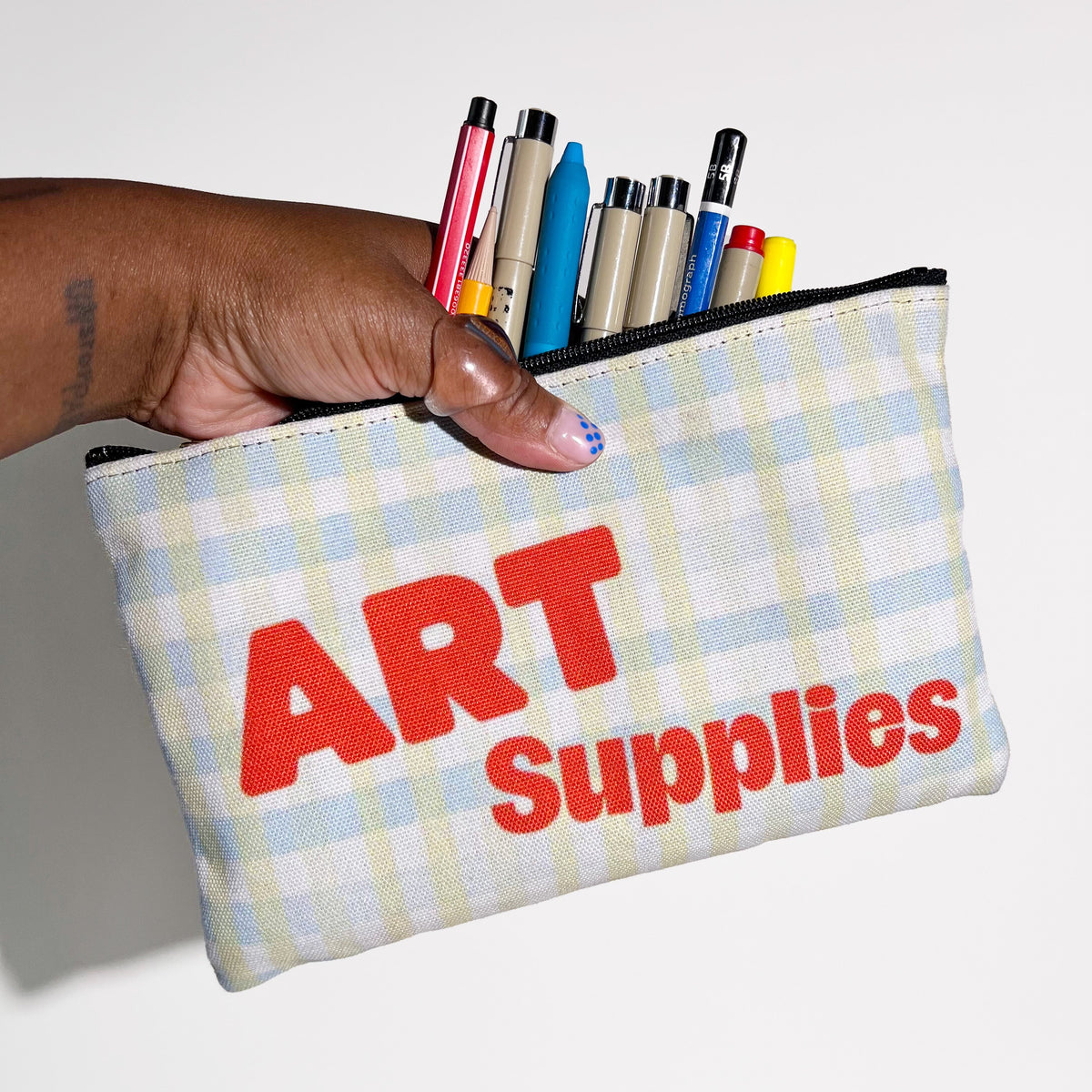 Two-sided polyester zip pouch with “Art Supplies” in a persimmon color with a white base and light blue and green gingham pattern. Includes a black zipper and gold zipper pull.