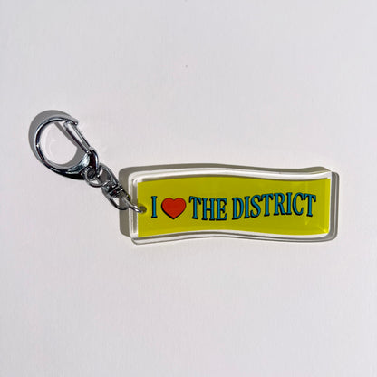 2.5” double-sided clear acrylic keychain with “I ♥ the District” in bright blue and red heart with wave typography treatment on green wave background. Silver D hook clasp and chain. DMV native DC Maryland Virginia bag charm for totes and crossbody bags. Washington DC hometown souvenir. 