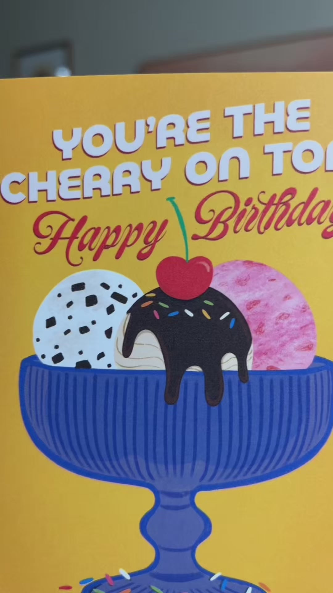 A2 size birthday card featuring blue antique glass ice cream bowl with chocolate chip, strawberry, and fudge salted caramel ice cream illustration on a warm yellow background. “You’re the cherry on top! Happy Birthday” text on the front in white and red. The message inside reads “Happy Birthday!”,. Envelope included.