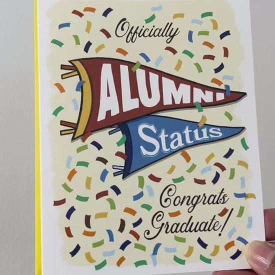 Video of A2 size vibrant graduation card featuring colorful confetti and collegiate pennant banners. “Officially Alumni Status, Congrats Graduate!” text on the front with "Alumni” on the top burgundy banner and “Status!" below on a blue banner. Colorful confetti and a message inside the card. Envelope included. 