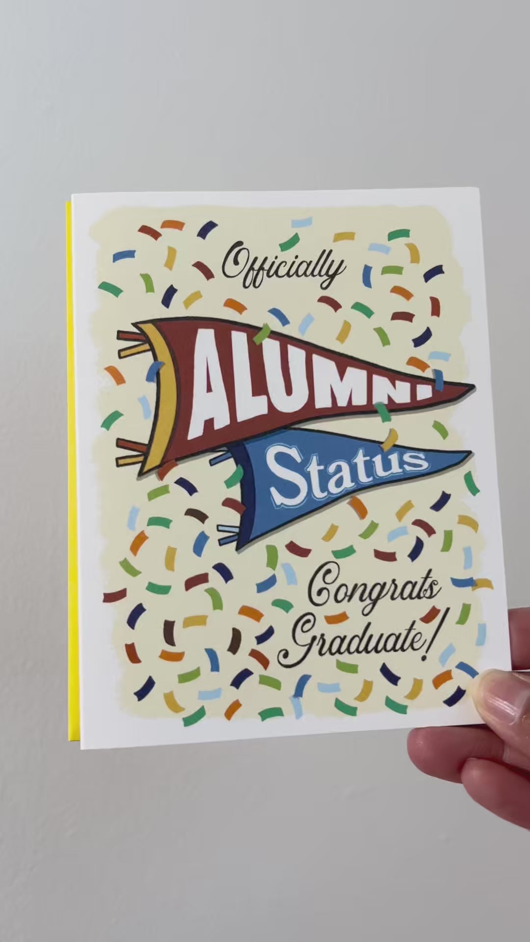 Video of A2 size vibrant graduation card featuring colorful confetti and collegiate pennant banners. “Officially Alumni Status, Congrats Graduate!” text on the front with "Alumni” on the top burgundy banner and “Status!" below on a blue banner. Colorful confetti and a message inside the card. Envelope included. 